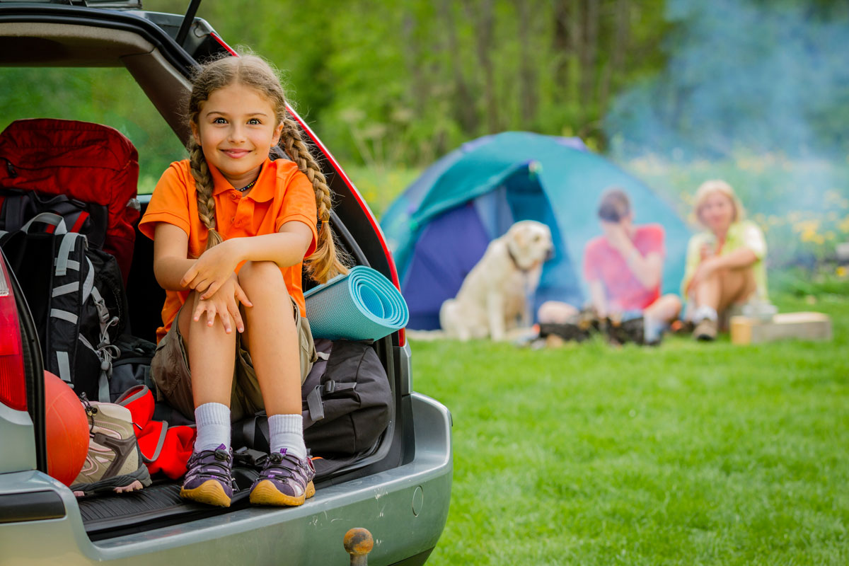 Kleines Mädchen sitzt hinten im Kofferraum mit Familie im Hintergrund im Zelt