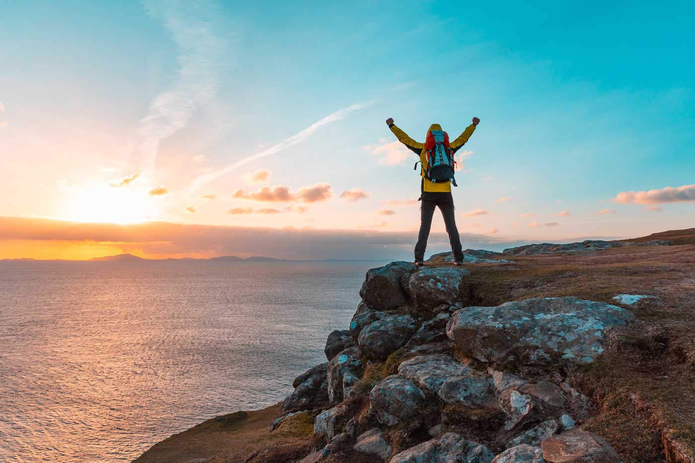 Glücklicher Wanderer mit gehobenen Armen auf der Spitze einer Klippe in Schottland bei Sonnenuntergang