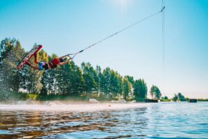 Junger Mann beim Wakeboarden auf einem See, macht Raley, Frontroll und springt über die Kicker und Slider.