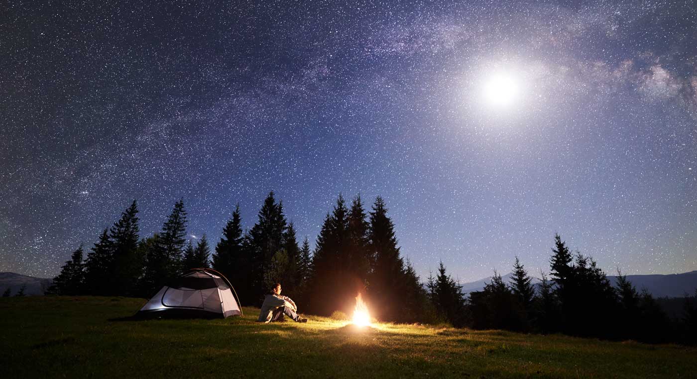 Männlicher Wanderer sitzt allein in der Nähe des Touristenzeltes am Lagerfeuer in einem grasbewachsenen Tal und genießt den nachtblauen Sternenhimmel mit Vollmond